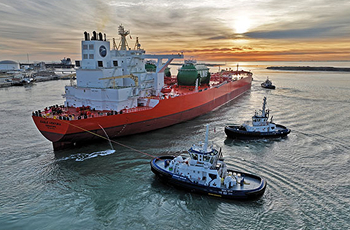 Large red cargo ship being guided by three tugboats in a harbor at sunset, with calm waters and a partly cloudy sky