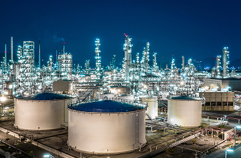Large industrial refinery at night with illuminated storage tanks and distillation towers under a dark blue sky