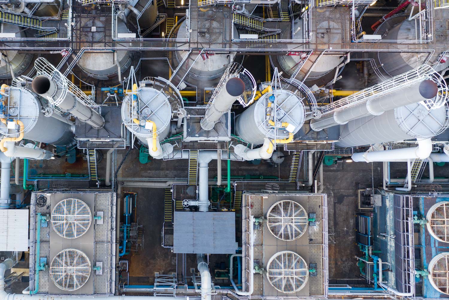 op-down view of an industrial facility showing large cylindrical tanks, metal pipes, cooling fans, and walkways