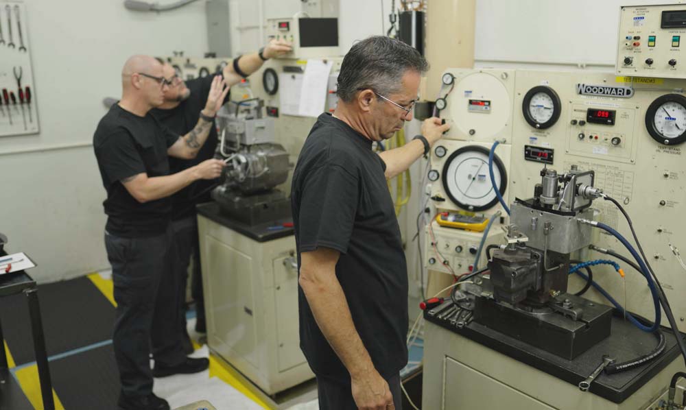 Technicians operating engine testing equipment and monitoring control panels in a workshop filled with tools and diagnostic instruments