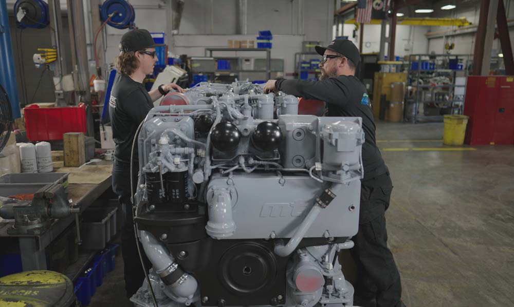 Two technicians working on a large industrial engine inside a spacious workshop, surrounded by tools and machinery