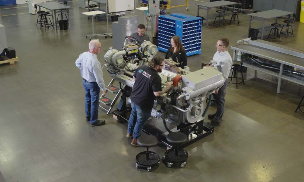 Group of people gathered around an industrial engine in a spacious workshop for a hands-on demonstration or training session, with tables and equipment in the background