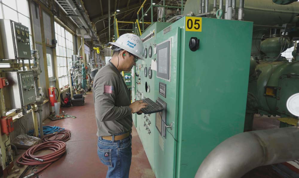 Engineer in a hard hat operating a green industrial control panel inside a large facility with machinery, equipment, and wheeled hoses visible