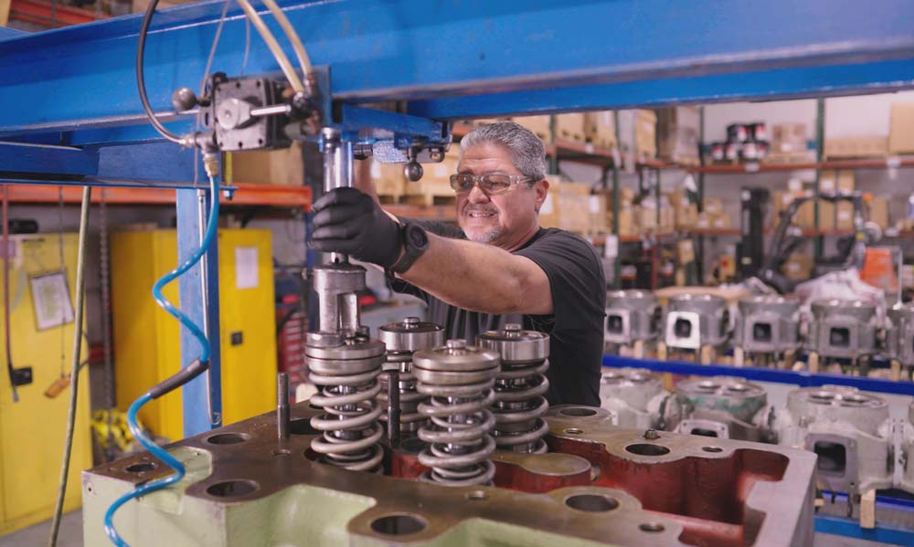 Technician using a specialized tool to assemble valve springs on an engine cylinder head in a workshop filled with equipment and engine parts